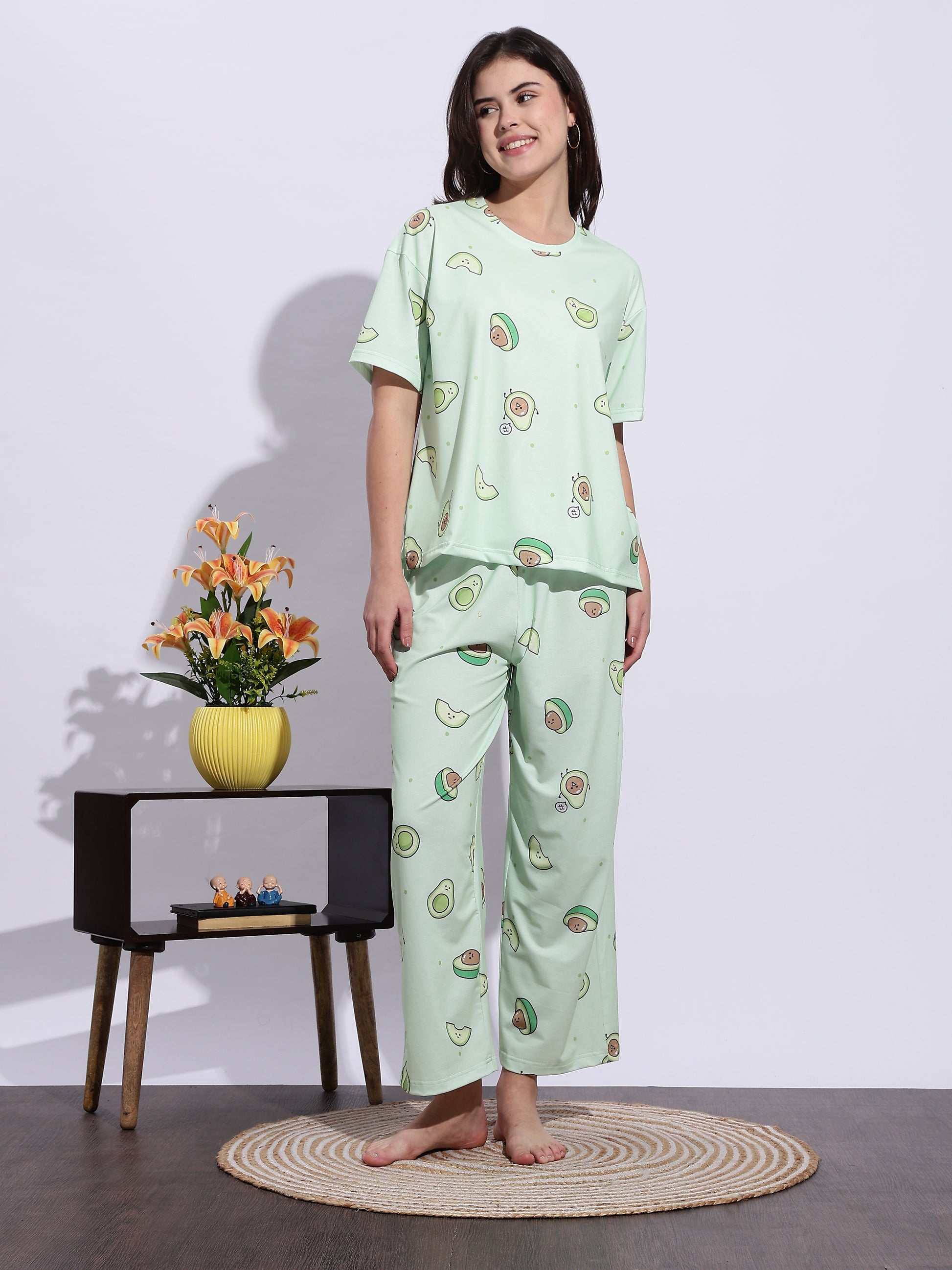 Woman wearing a light green pajama set with fruit patterns, standing in a room with a small table and flowers.