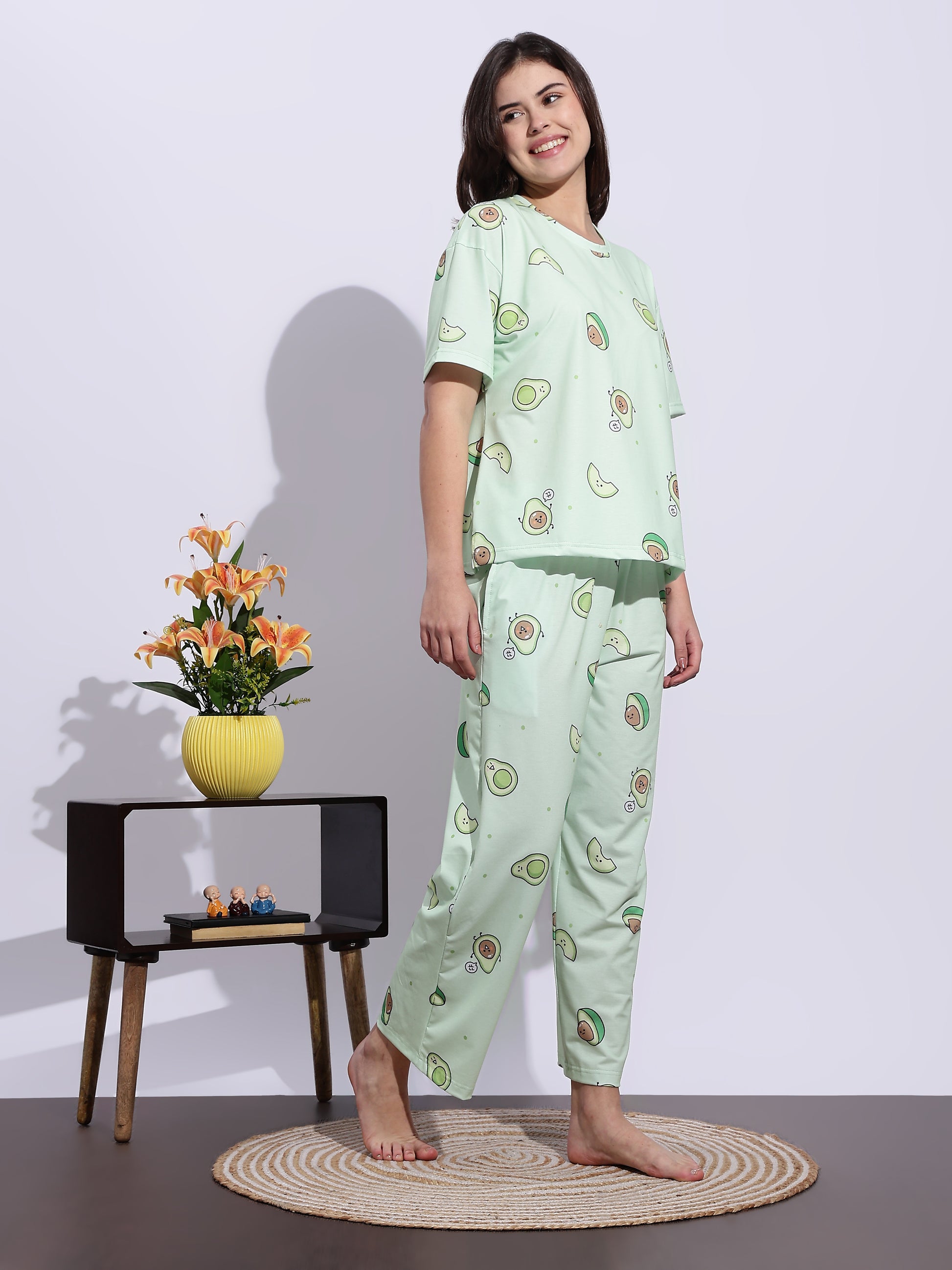 Woman wearing a green pajama set with avocado pattern standing in a room with a small table and flowers.