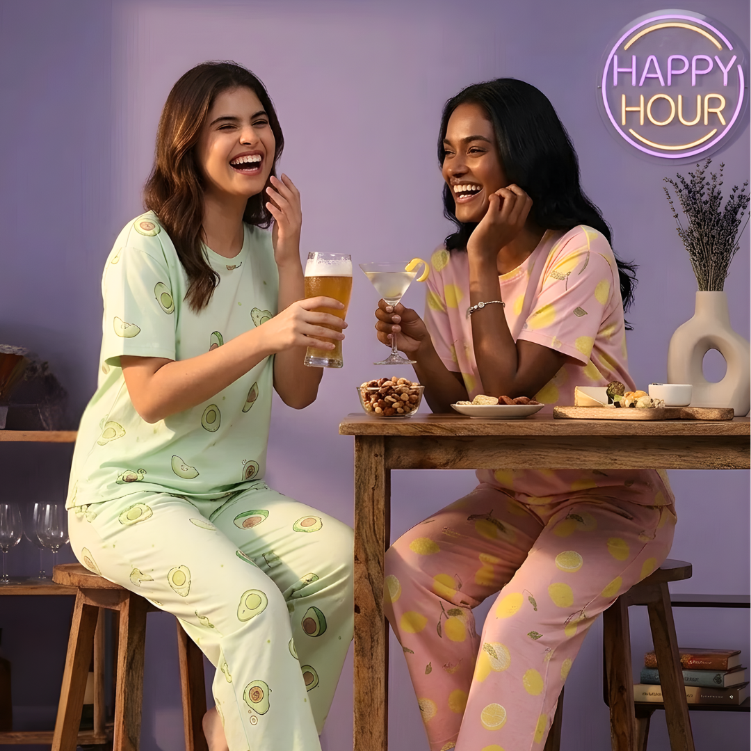 Two women in pajamas enjoying drinks and snacks at a table with 'Happy Hour' branding.